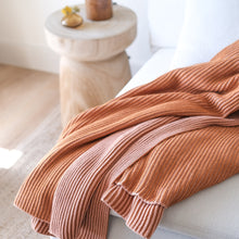 Load image into Gallery viewer, Burnt orange and pink clay ribbed cotton throw blanket draped over a cream-colored sofa with natural wood side table in background