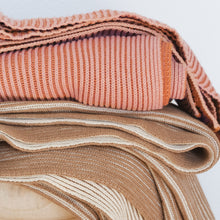 Load image into Gallery viewer, Stacked ribbed knit cotton throw blankets in burnt orange with pink clay and cacao with oat milk colorways on a wooden stool
