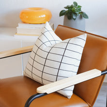 Load image into Gallery viewer, Modern grid cotton knit pillow in medium black grid pattern styled on camel-toned armchair with book and mug
