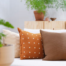 Load image into Gallery viewer, Burnt orange geometric pillow with mini checker pattern styled alongside a neutral ribbed pillow and textured cream cushion, set against a cozy natural wood and greenery backdrop.
