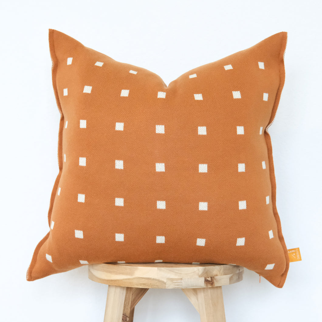 Burnt orange pillow cover with small white square pattern, styled on a light wood stool.