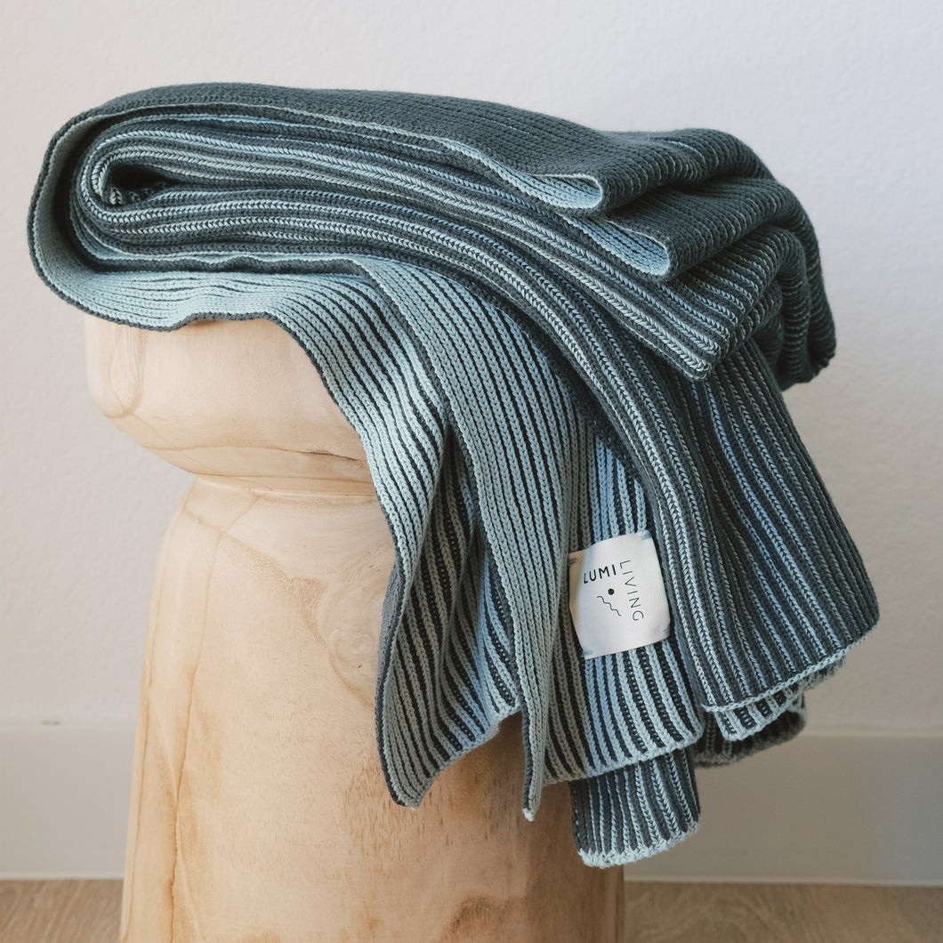 Folded ribbed cotton knit throw blanket in mint blue and dark green on a carved wooden stool with Lumi Living tag visible